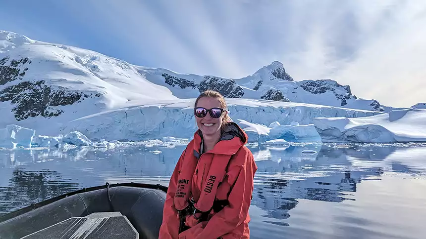 Female in red parka and life vest sits on nose of inflatable skiff with blue sky, snow covered mountains and icebergs behind