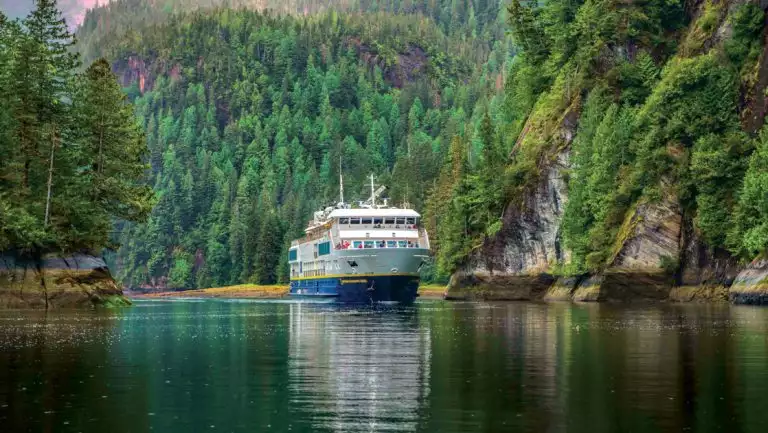 Nat Geo Quest small ship with blue hull & white upper decks cruises on glassy water between lush green mountains.