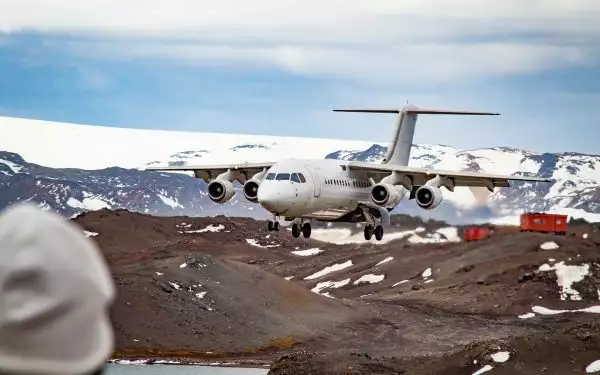 White airplane descends to a landing strip in Antarctica surrounded by jagged white mountains and brown dirt ground,