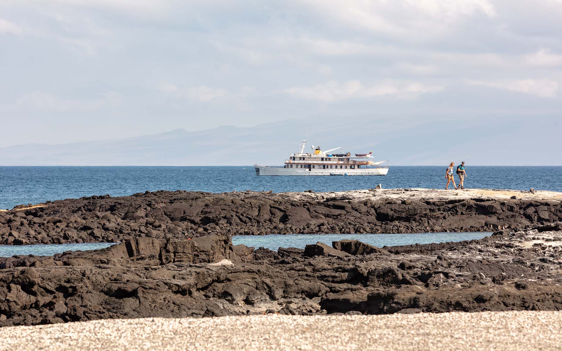 A small white Galapagos cruise ship seen with the foreground of lava rock and two travelers hiking on the shore in front of the ship