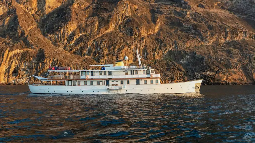 Grace boat in Galapagos with white hull & 2 upper decks, wood finishes & many windows, sits beside golden cliffs in calm sea.