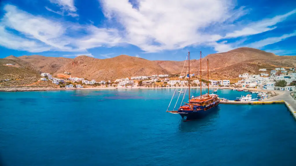 Aerial view of small motorsailor with blue hull & teak decks docked in teal sea at a small island town in Greece's Cyclades.
