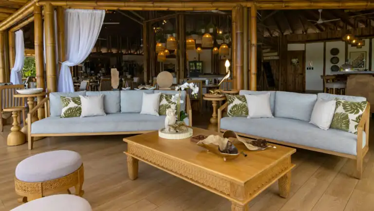 Open-air lounge at La Selva Ecuador with thatch roof, wood floor & coffee table & padded blue couches beside a lake.