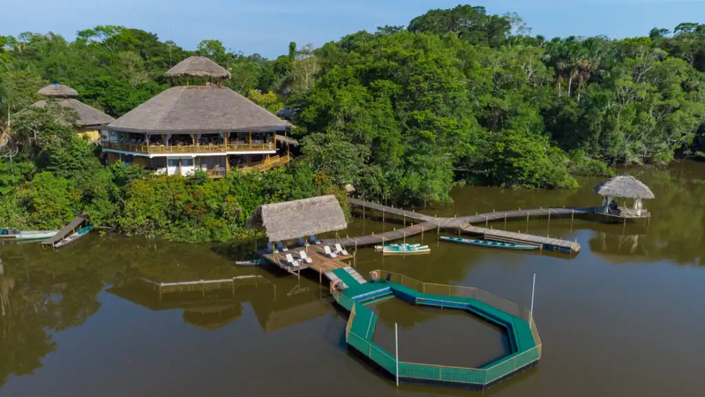 Aerial view of La Selva Lodge with hexagonal thatch-roof buildings beside a lake with 4-way wood dock & green-circled natural pool.