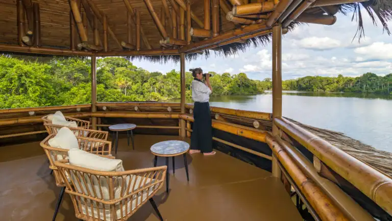 Woman stands iby padded white chairs & small tables in thatch-roof observation tower & uses binoculars to view the lake below.