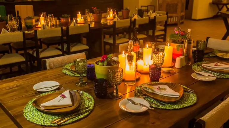 A beautifully decorated dining table with candles, flowers, and colorful glassware. The table is set with plates, napkins, and silverware, ready for a meal. The room is dimly lit with warm lighting, creating a cozy and inviting atmosphere.