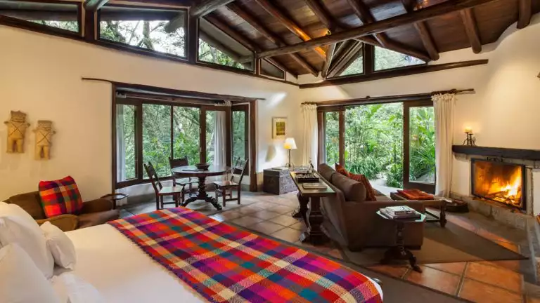 Guest room in small machu pichu hotel with white walls and large wooden beams, a king bed with white sheets and a colorful blanket, a fireplace and seating area that opens up to the outdoors