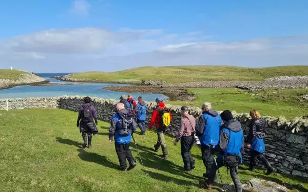 Small group of travelers on a Northern European tour walk over grass plains beside small inlet under blue skies.