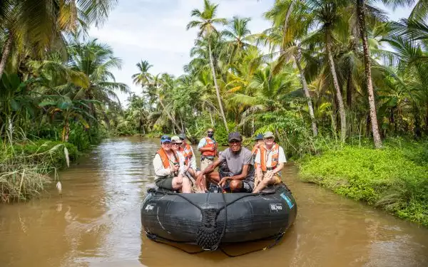 Black Zodiac boat with Panama small ship cruise travelers floats on muddy river under swaying palm trees.