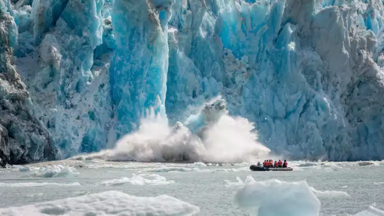 Zodiac dinghy with Wild Alaska Escape cruise travelers motors past an icy blue glacier as it calves & creates a large splash.