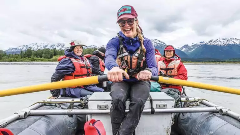 Young woman smiles while rowing a boat full of Wild Alaska Escape travelers down the Chilkat River with mountains behind.