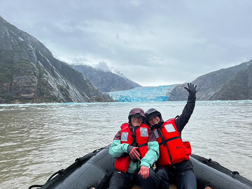 A mother and adult daughter in red life jackets pose on the bow of a Zodiac in front of a bright blue glacier in Alaska