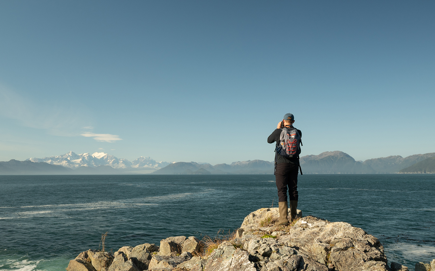 A man in boots stands on rocks facing away from the camera looking out to the ocean with snowcapped mountains