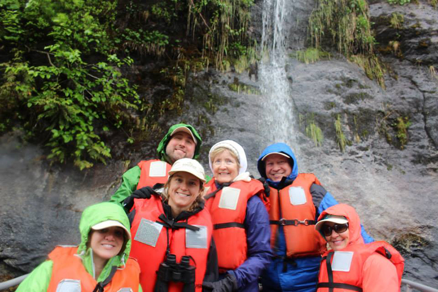 A multigenerational family huddles near a waterfall while in a zodiac wearing red lifejackets