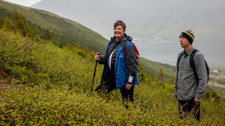 Woman hiker in blue jacket smiles as man in gray shirt looks uphill on a hike in green shrubs along Iceland's Westfjords & North Coast.