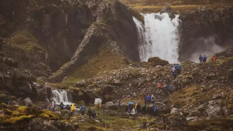 Travelers on Iceland's Westfjords & North Coast cruise hike over tundra & boulders near large waterfall in misty conditions.