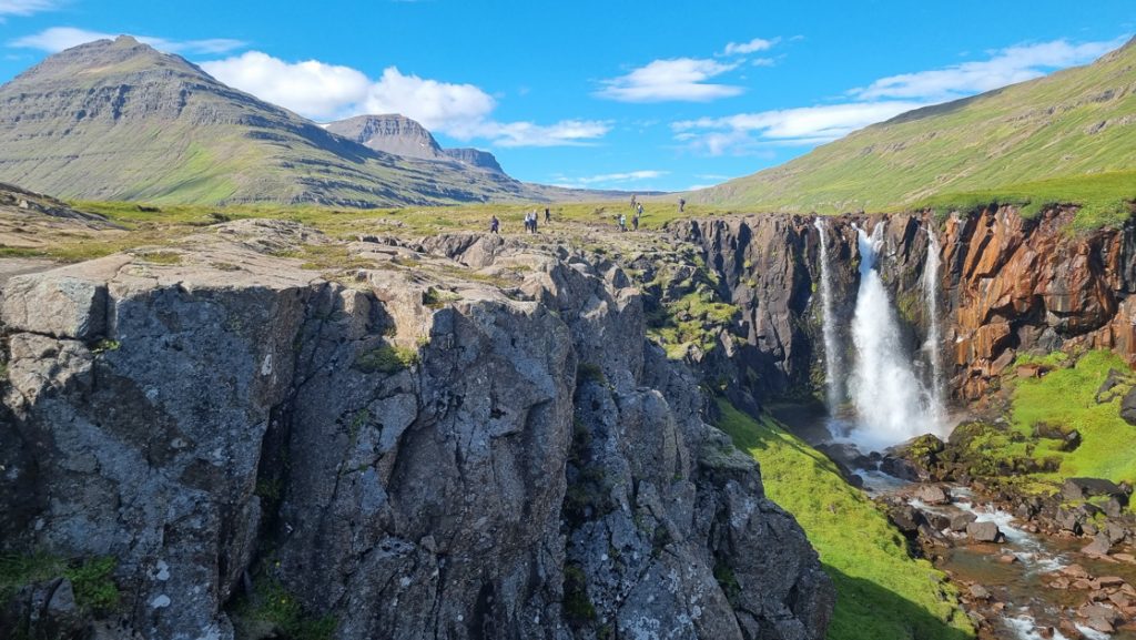 Hikers walk back from large cliffs overlooking gushing waterfall under sunny skies & among green grass in Iceland.
