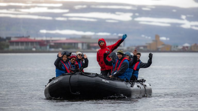 Zodiac boat with waving Iceland's Westfjords & North Coast cruise guests moves through calm water under cloudy skies.