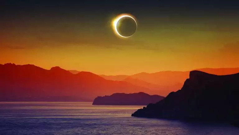 Full solar eclipse with moon blocking sun over arctic landscape of mountains & sea in orange, red, purple & gold.