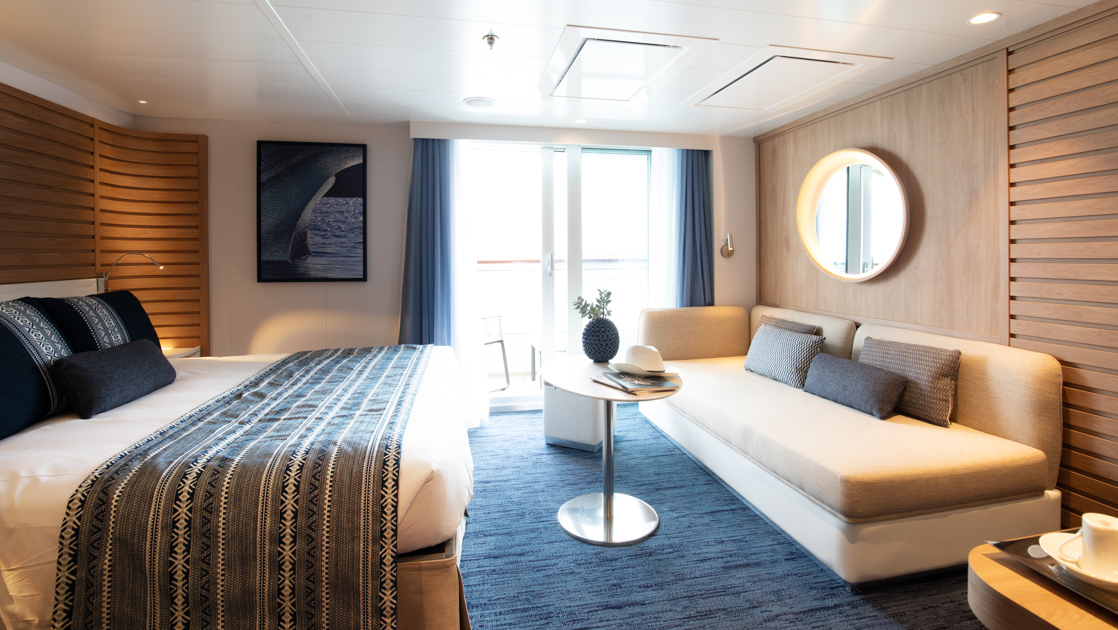 Deluxe Suite aboard Le Laperouse French luxury ship, with king bed, small table, couch, private balcony & ethic-chic decor.