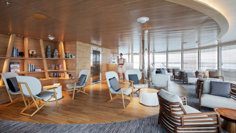 Panoramic lounge on Le Laperouse ship with floor-to-ceiling windows, light wood, art, bookshelves & padded chairs.
