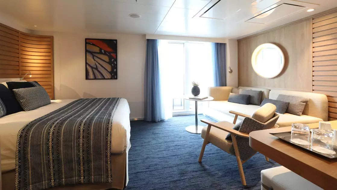 Privilege Suite aboard luxury ship Le Laperouse, with king bed, couch, table, chair & private balcony, plus natural decor.