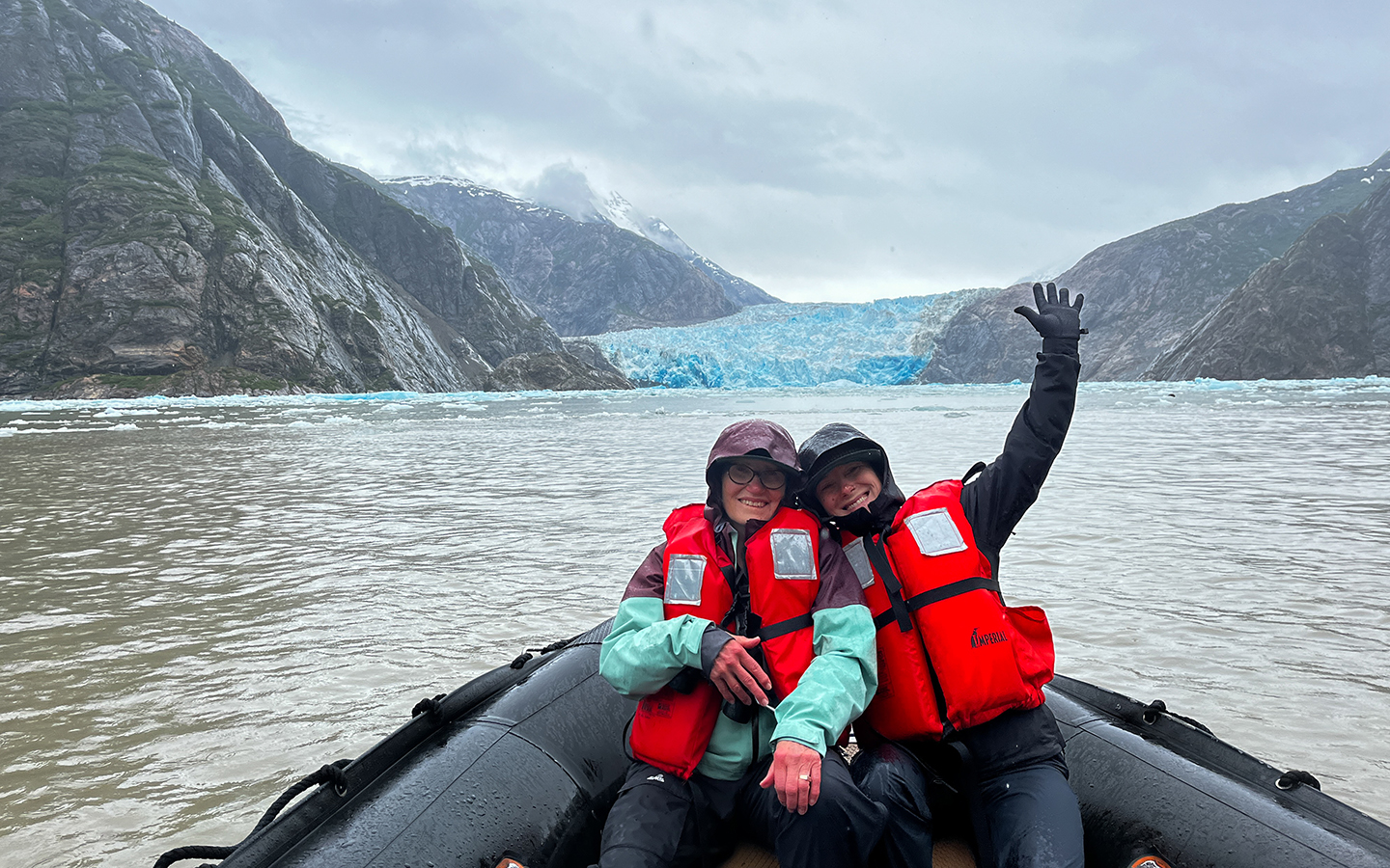 A mother and adult daughter sit on the bow of Zodiac with one arm up in red life jackets in front of a bright blue glacier in Alaska