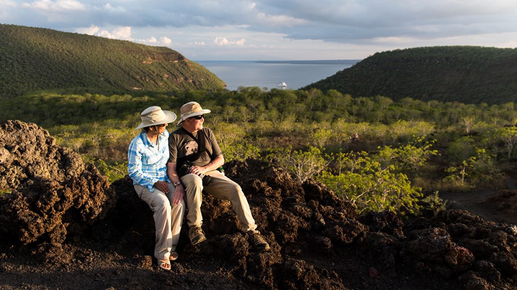 A male and female traveler sit atop lava rock at Tagus Cove in the Galapagos Islands with green trees and the ocean in the distance below them.