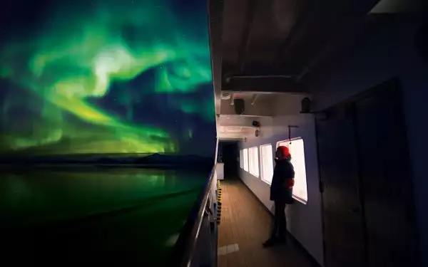 Guest aboard small ship wearing red beanie looks up at the sky at the bright green ribbons of light, aka northern lights.