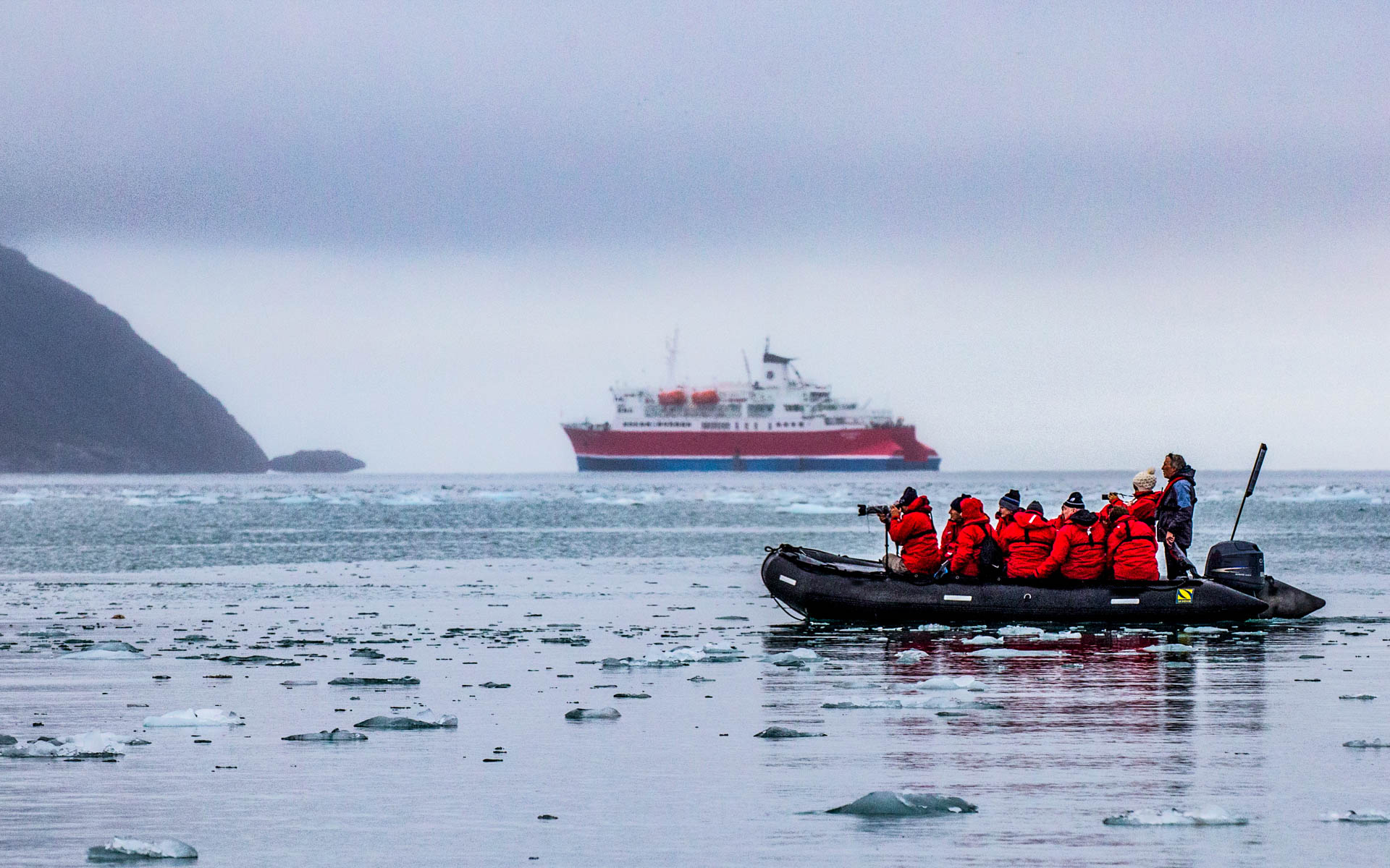 Arctic cruise travelers in red jackets take photographs of iceberg bits in the water on a Zodiac, with a small red polar ship in the background.