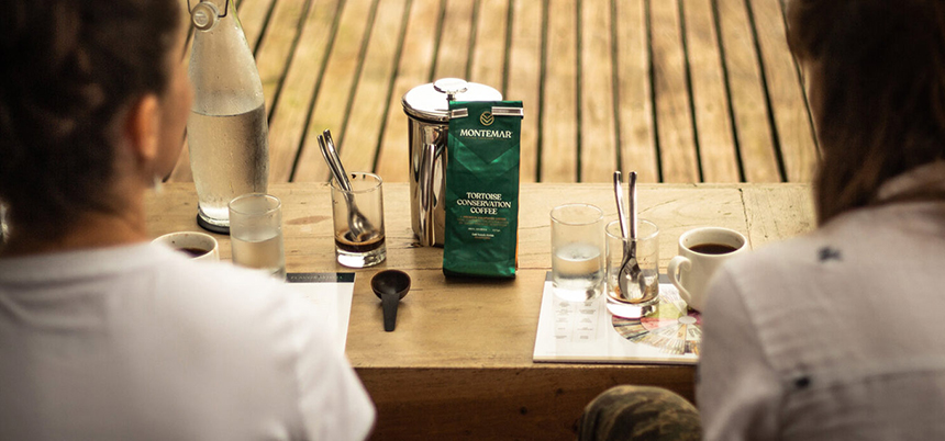 A green Turtle Conservation Coffee bag seen on a table in front of two travelers