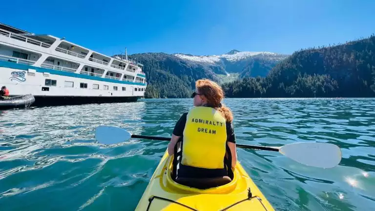 Woman with red hair in yellow PFD & tandem kayak boat sits holding paddle by blue & white small ship on a sunny day in Alaska.