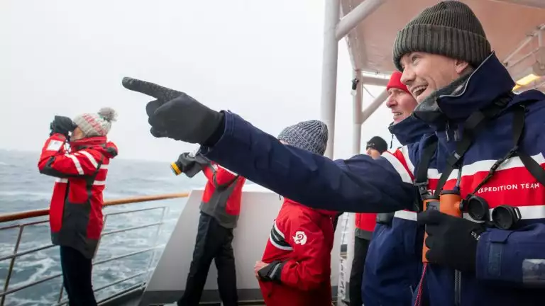 Explorers dressed for the cold weather in gloves and hats look out off the stern of a small cruise ship