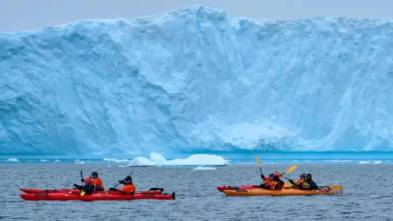 cold weather Kayakers navigate icy waters in front of massive iceberg thats light blue in color