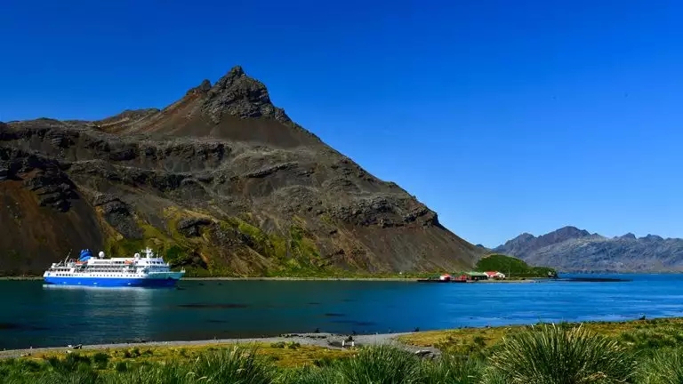 Small cruise ship sits in the bay of blue waters surrounded by steep mountains on a blue sky day