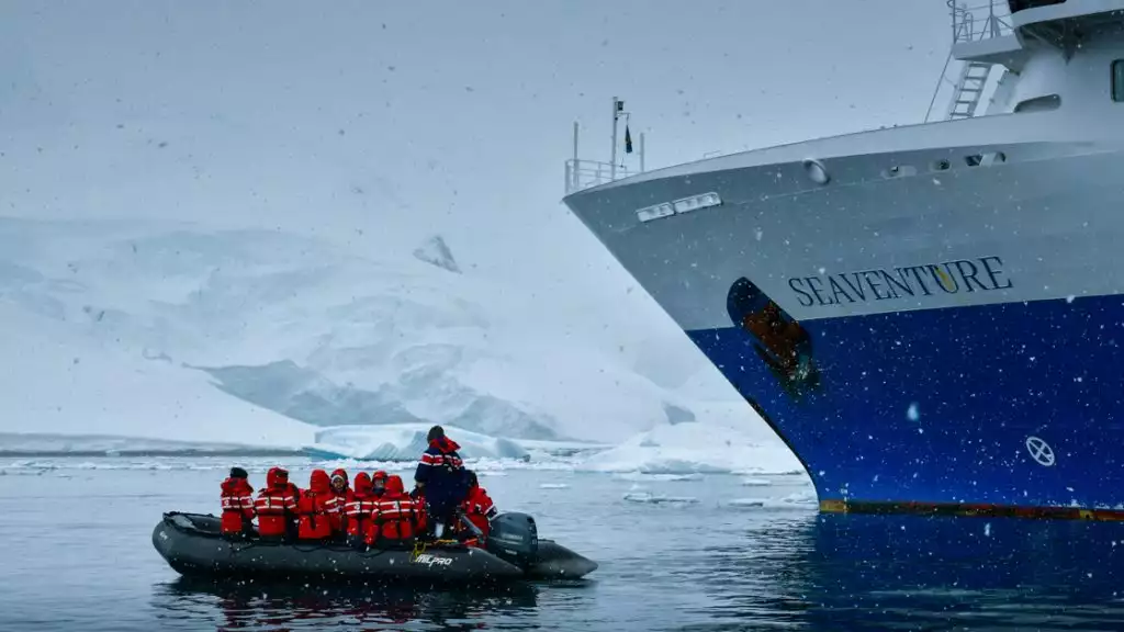 Small skiff filled with travelers in red winter coats ventures off the small cruise ship into the snowy landscape and snowy mountains