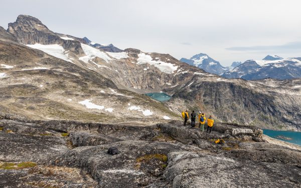 A group of five travelers in yellow jackets stands on a rocky ridge overlooking fjords below and mountains dotted with snow on the ridge above them