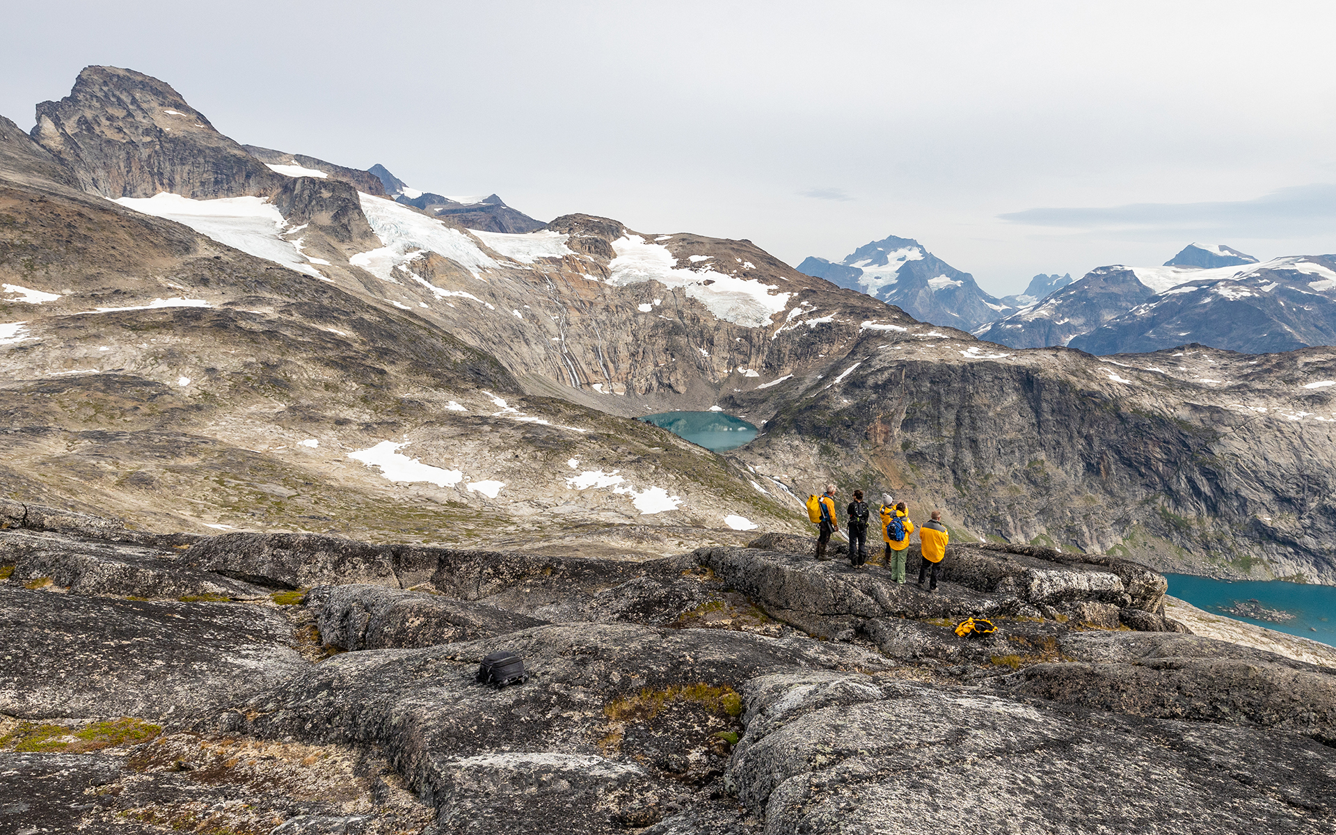 A group of five travelers in yellow jackets stands on a rocky ridge overlooking fjords below and mountains dotted with snow on the ridge above them