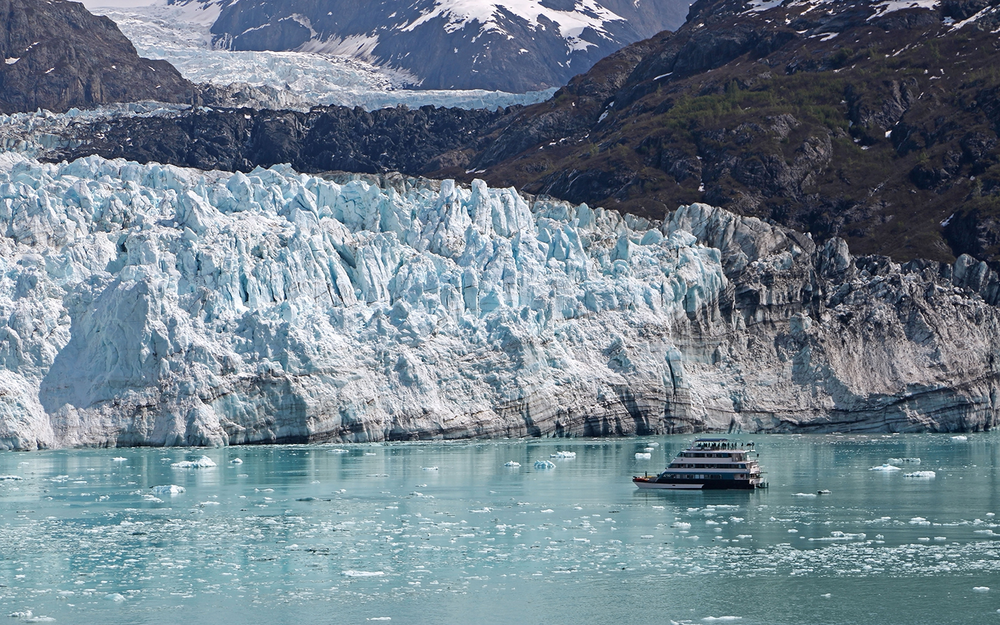 A small ship is seen alone and dwarfed in front of the blue and white Margerie Glacier at Glacier Bay National Park, Alaska