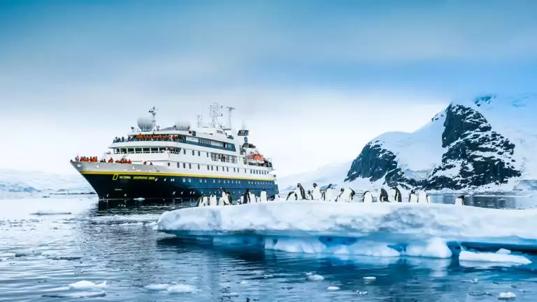 National Geographic Orion small ship with blue hull & white upper decks sits in calm sea by small iceberg with penguins.