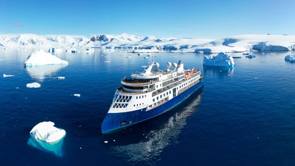 Aerial view of Ocean Explorer expedition ship in icy water with blue hull & white upper decks, slim bow, many windows & teak decks.