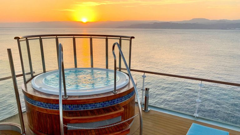 Round Jacuzzi with glass panels on one corner & wood & tile exterior sits atop teak deck of Ocean Explorer ship at sunset.