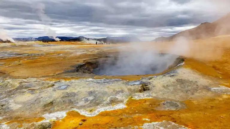 Geothermal pool with smoke rising out of it among an orange & tan landscape under cloudy skies in Iceland.