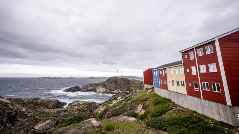 Row of red, yellow & blue buildings beside rocky seaside hills under cloudy skies, seen on a southern Greenland cruise.