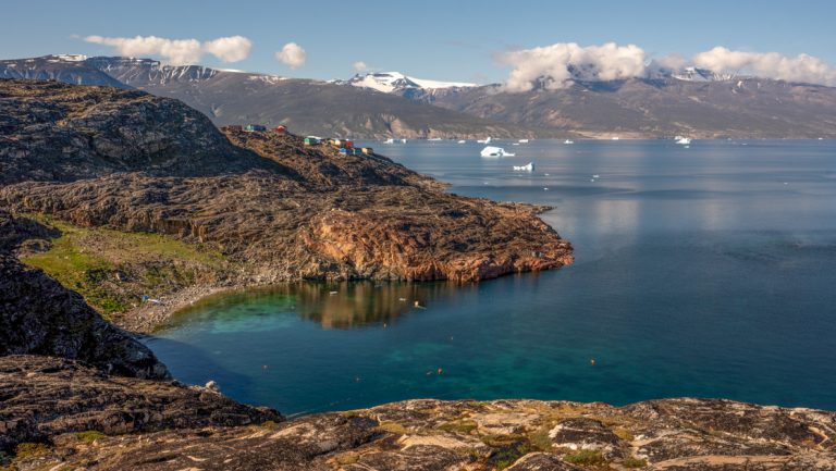 View onto adjoining coves with green hills, tan rocks & small colorful homes, seen on a southern Greenland cruise.