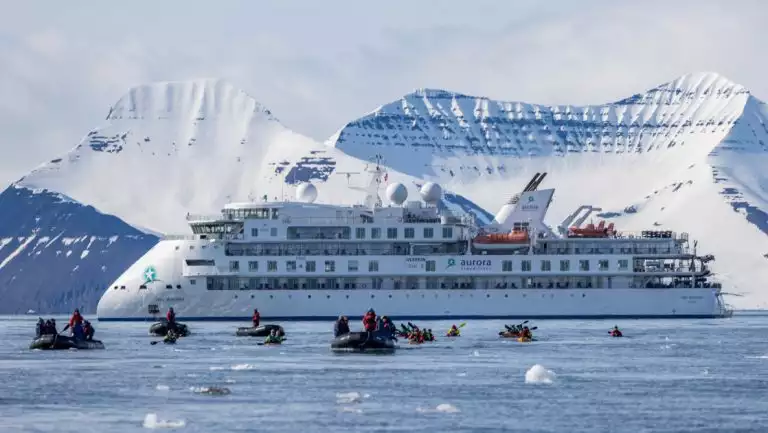 White expedition ship Greg Mortimer sits in calm icy water by snow-covered peaks as travelers kayak & Zodiac cruise nearby.