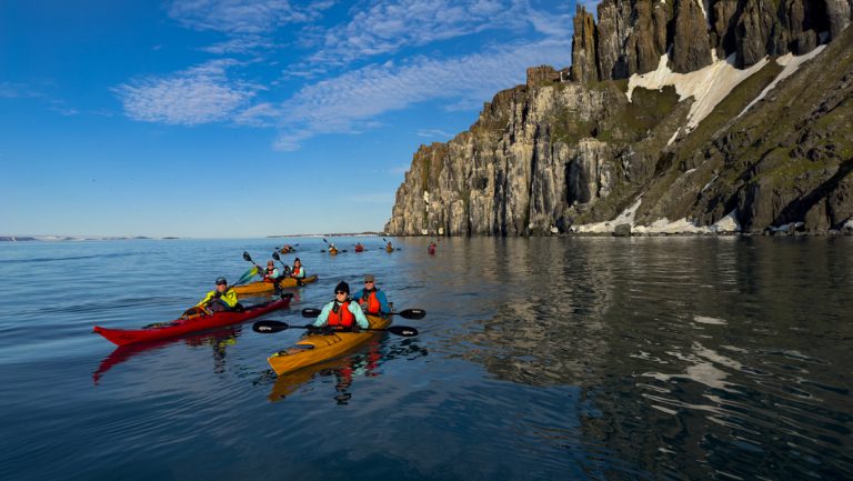 Tandem kayakers in red & yellow boats sit atop glassy water beside tall, rocky cliffs on a sunny day in Spitsbergen.