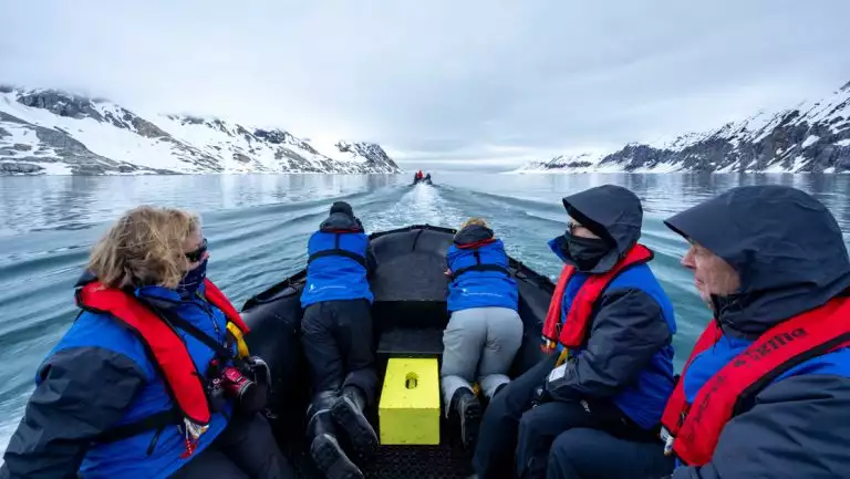 Spitsbergen: Realm of the Ice Bear Cruise guests in red life jackets & blue coats sit in Zodiac & cruise among snowy peaks.