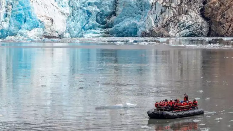 Rigid DIB dinghy with Wild Alaska Odyssey cruise travelers in orange PFDs motors near a large blue glacier.