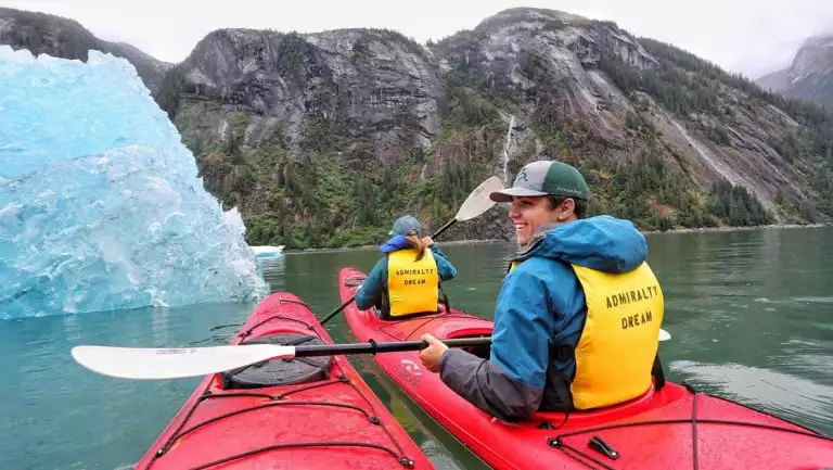 Young man & woman sit in red tandem kayak holding paddles in front of blue glacier bergy bit in Alaska.